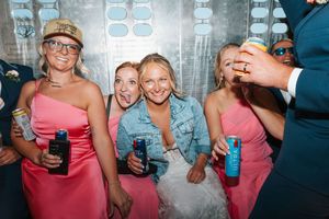 Bridesmaids in pink dresses laugh and hold drinks in a photo booth at a wedding reception.