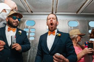 Groomsmen in navy suits and orange bow ties celebrate energetically at a lively wedding reception party.