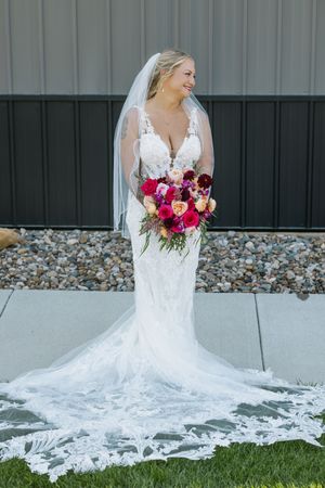 Elegant bride in fitted lace gown with long train holds vibrant pink and coral bouquet outside gray barn venue.