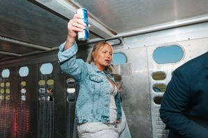 Blonde woman in denim jacket raises beer can in celebration inside a party vehicle.