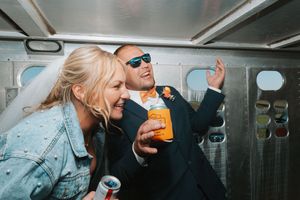 Bride and groom laughing together in a vintage vehicle, groom wearing sunglasses and holding an orange drink on their wedding day.