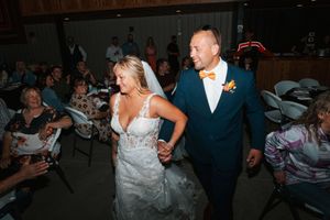 Smiling bride and groom in navy suit with orange bow tie walk hand in hand through wedding reception crowd.