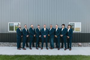 Nine groomsmen in matching teal suits pose in a row outdoors against a modern gray barn wall.