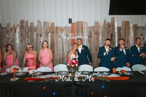 Wedding party poses at head table with Mrs. & Mr. sign, bridesmaids in pink and groomsmen in navy suits.