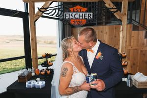 Bride and groom kissing under Fischer Est. 2025 sign at rustic barn wedding reception dessert table.
