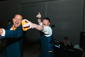 Two groomsmen celebrate energetically on wedding reception dance floor holding drinks.
