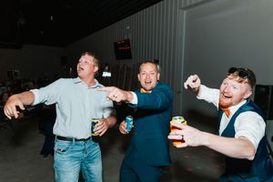 Three men celebrating and dancing energetically at wedding reception inside venue holding drinks.