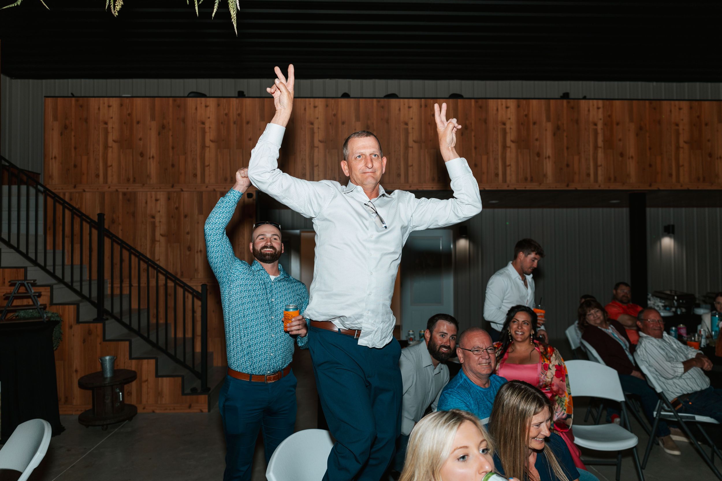 Man celebrates with arms raised at a lively wedding reception venue with guests seated around him.