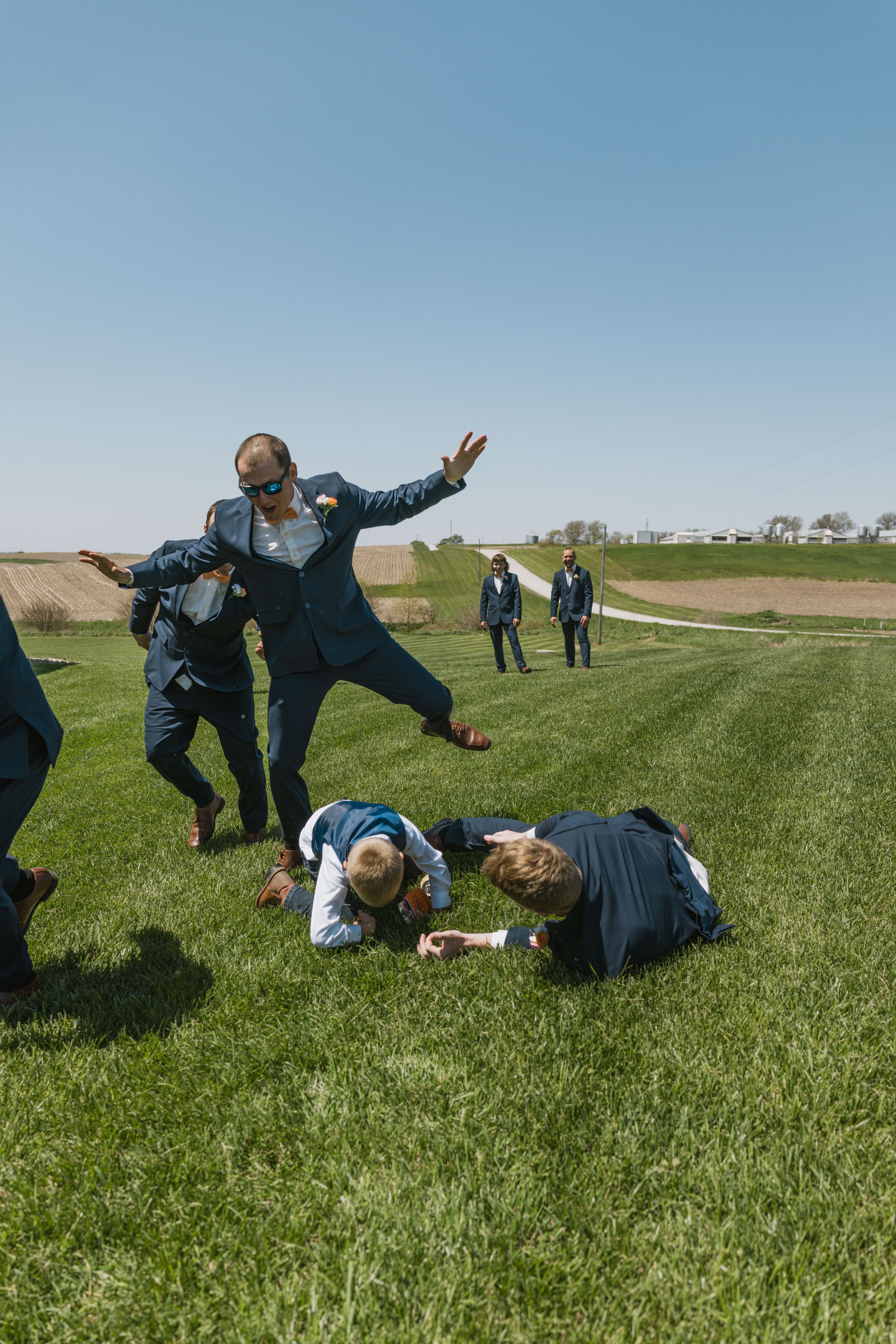 Groomsmen in blue suits playing on green grass field outdoors on a sunny day at a wedding celebration.