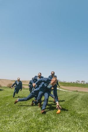 Groomsmen in navy suits playing football on green grass field under clear blue sky at wedding.
