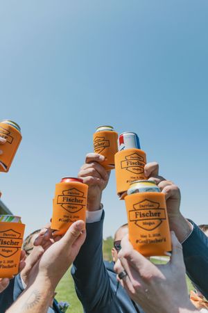 Wedding party raises custom orange Fischer wedding koozie cans in a celebratory toast under a bright blue sky.