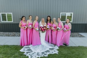 Bride in white gown with six bridesmaids in pink dresses holding bouquets posing outdoors at wedding venue.