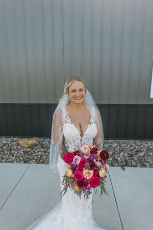 Smiling bride in lace wedding gown and veil holding vibrant pink and peach bouquet outdoors.