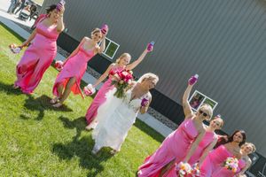 Bride in white gown with bridesmaids in pink dresses celebrating outdoors, raising bouquets joyfully on a sunny wedding day.