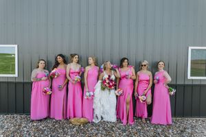 Bride in white gown with bridesmaids in pink dresses holding bouquets, posing in front of a gray barn.