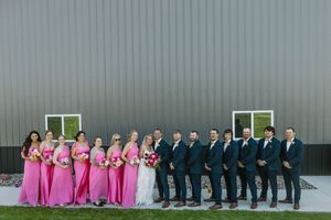 Wedding party poses outside gray barn with bridesmaids in pink dresses and groomsmen in navy suits holding bouquets.