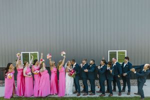Wedding party posing outside barn, bridesmaids in pink dresses and groomsmen in navy suits raising bouquets.