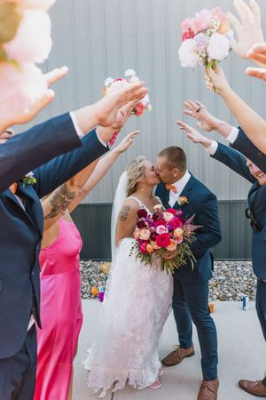 Bride and groom share a kiss under an arch of raised hands and colorful bouquets during their wedding ceremony.