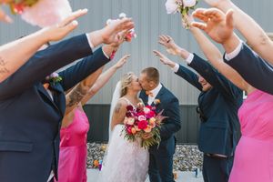 Bride and groom kissing under arch of raised hands formed by wedding party in pink and navy attire.