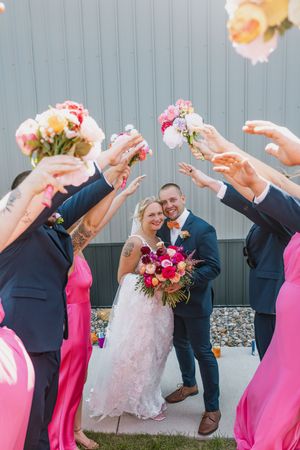 Bride and groom walk through arch of raised floral bouquets held by wedding party in pink and navy.