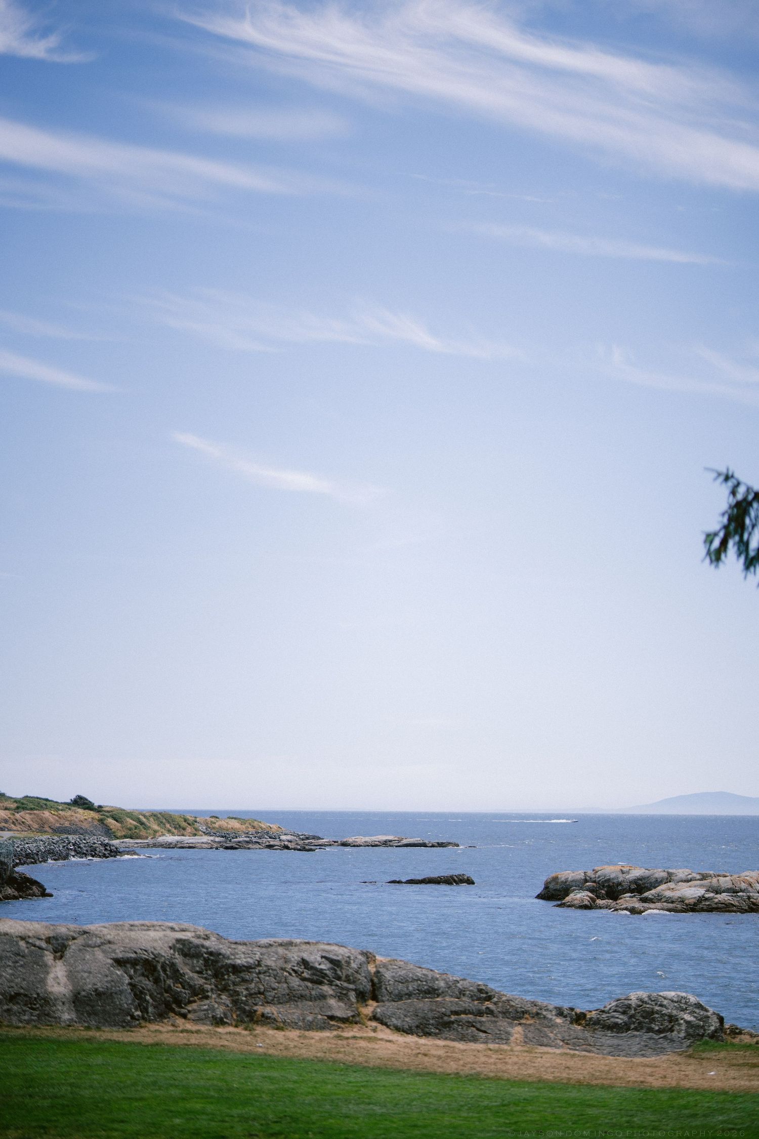Rocky coastal shoreline with calm blue ocean waters under a clear sky with wispy clouds.