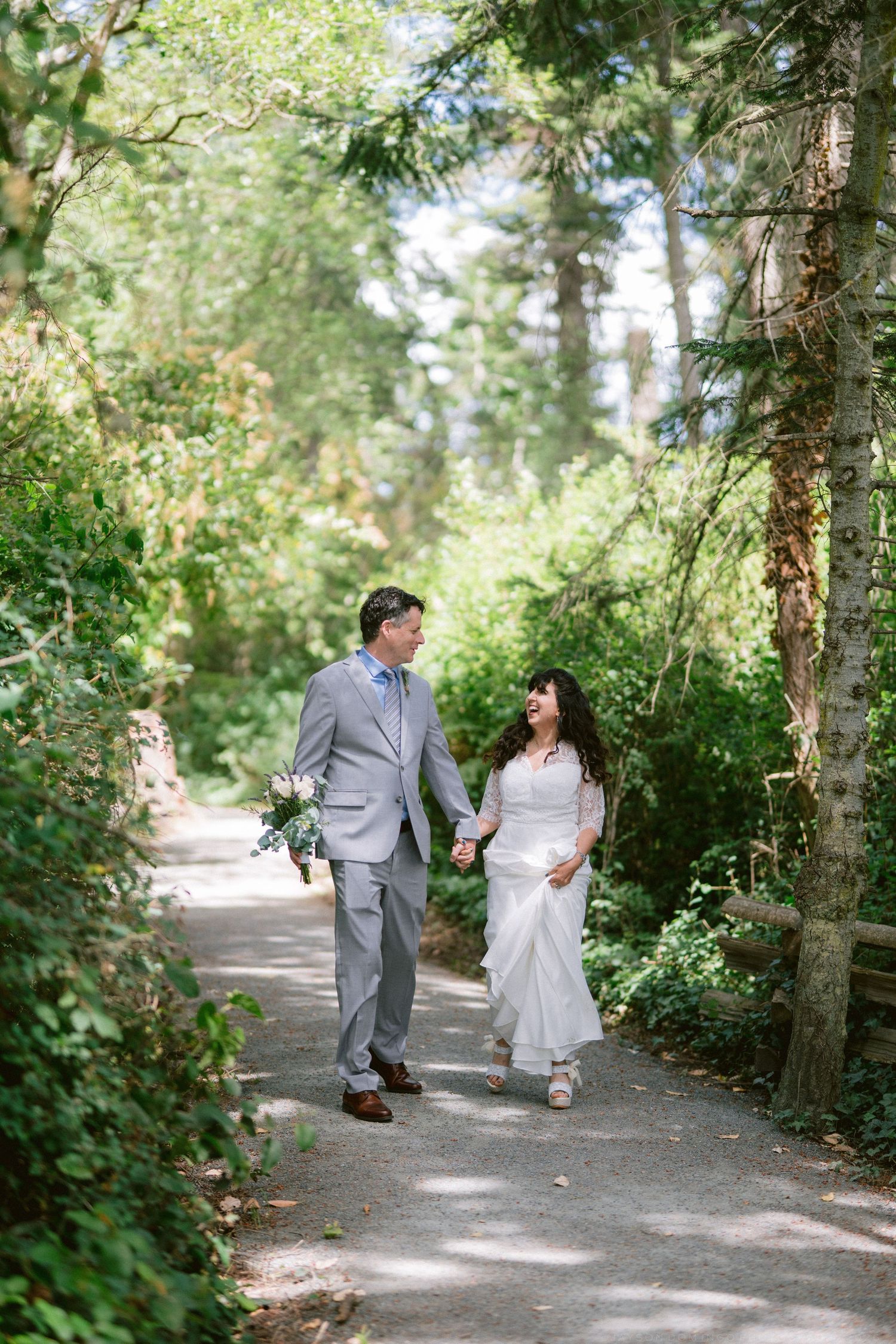 Bride and groom walking hand in hand along a sunlit tree-lined path on their wedding day.