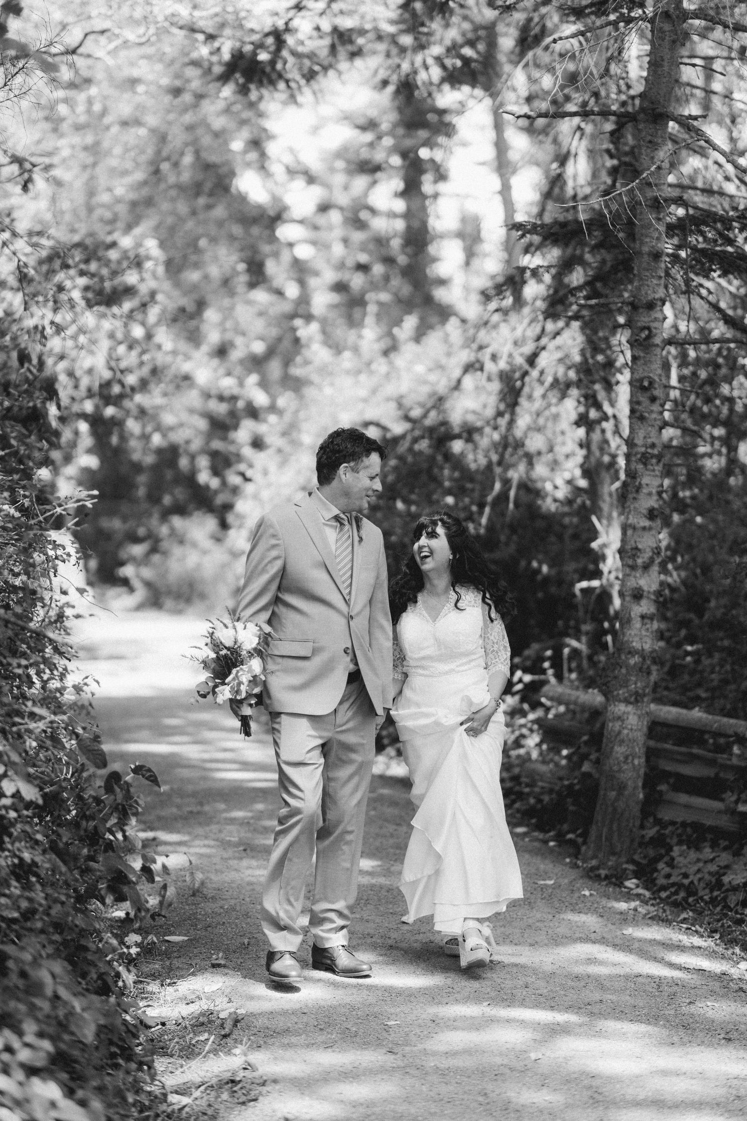 Black and white photo of a bride and groom walking hand in hand along a sunlit forest path.