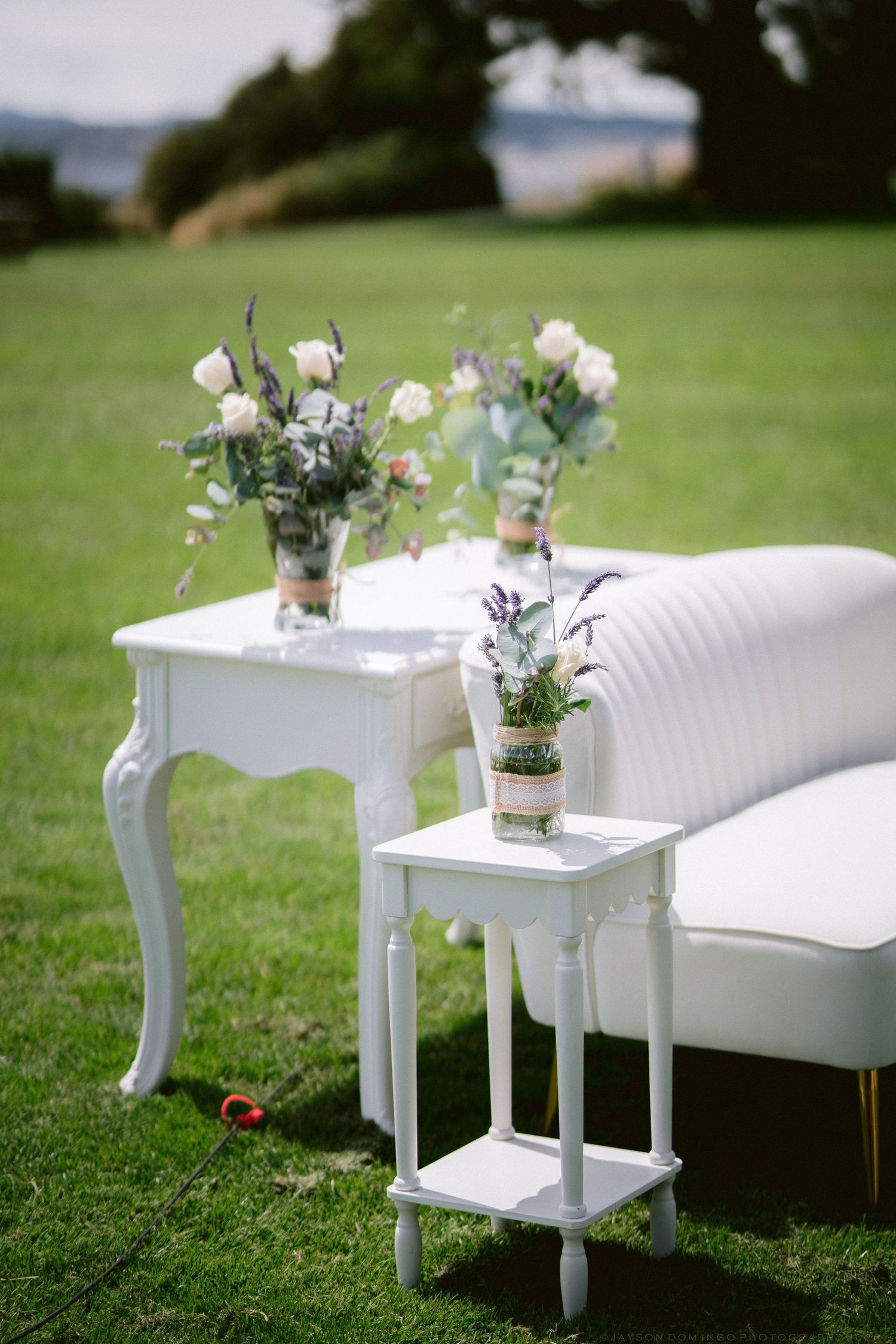 Elegant white outdoor wedding setup with floral arrangements on white furniture on green lawn.