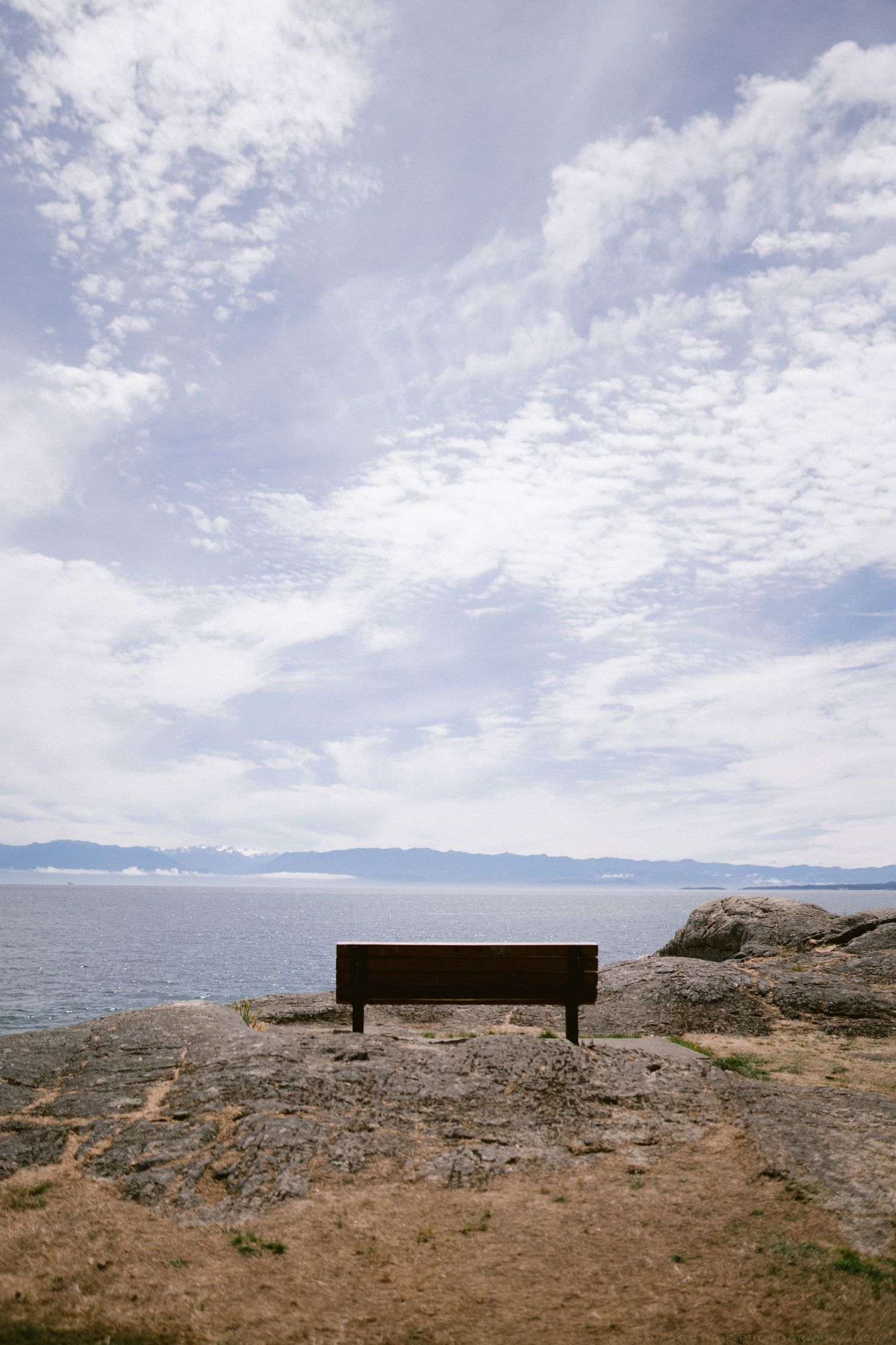 Lone wooden bench on rocky coastal cliff overlooking calm blue ocean under dramatic cloudy sky.