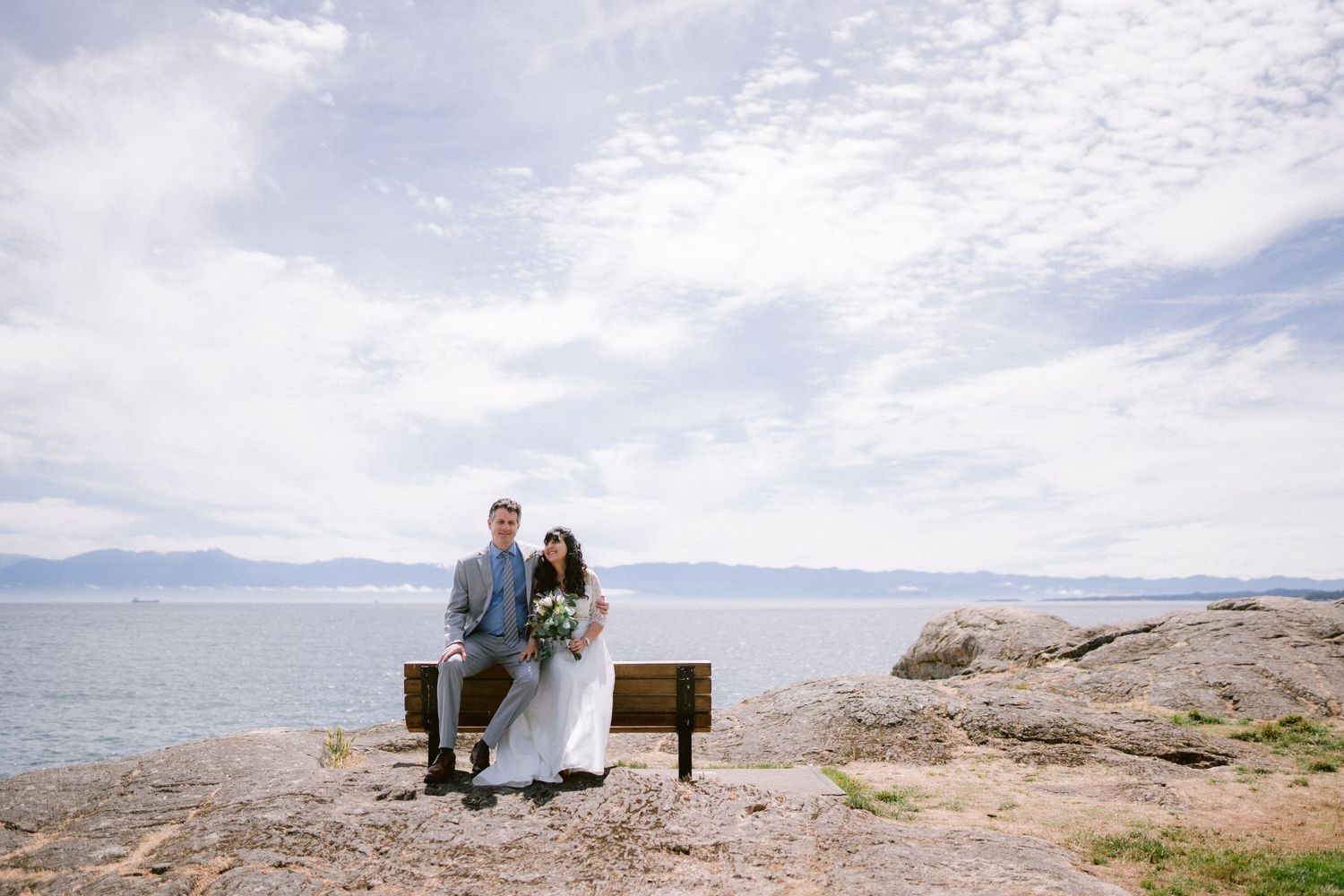 Bride and groom sitting together on wooden bench with rocky coastline and cloudy sky behind.