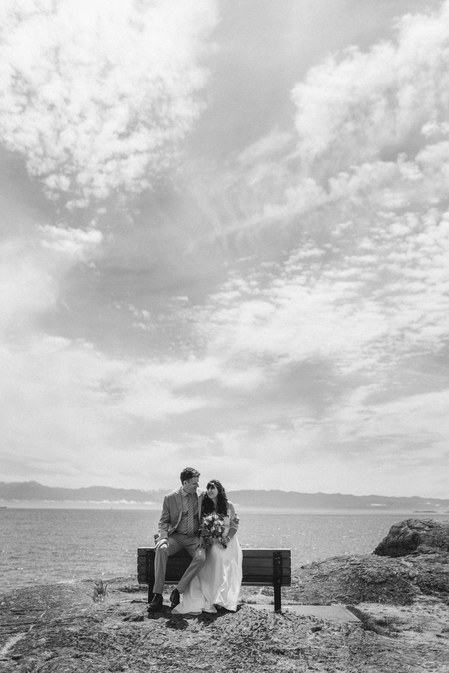 Black and white photo of bride and groom sitting on bench with dramatic ocean sky backdrop.