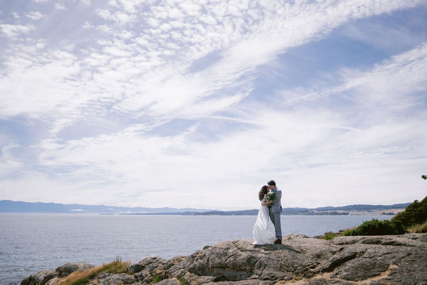 A bride and groom share a kiss on rocky coastal cliffs with a vast ocean and cloudy sky behind them.