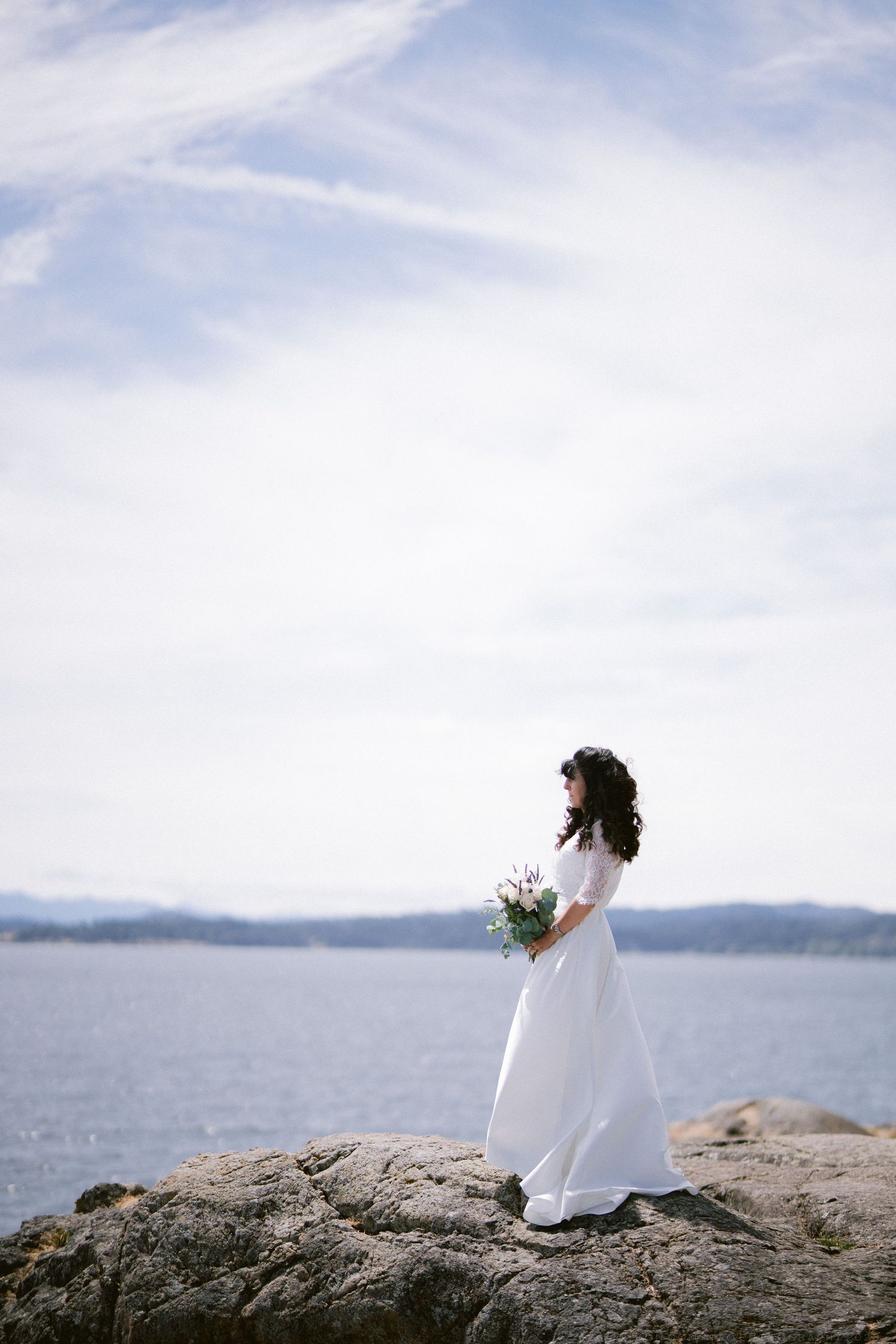 Bride in white gown holding bouquet stands on rocky shore with ocean and mountains in background.