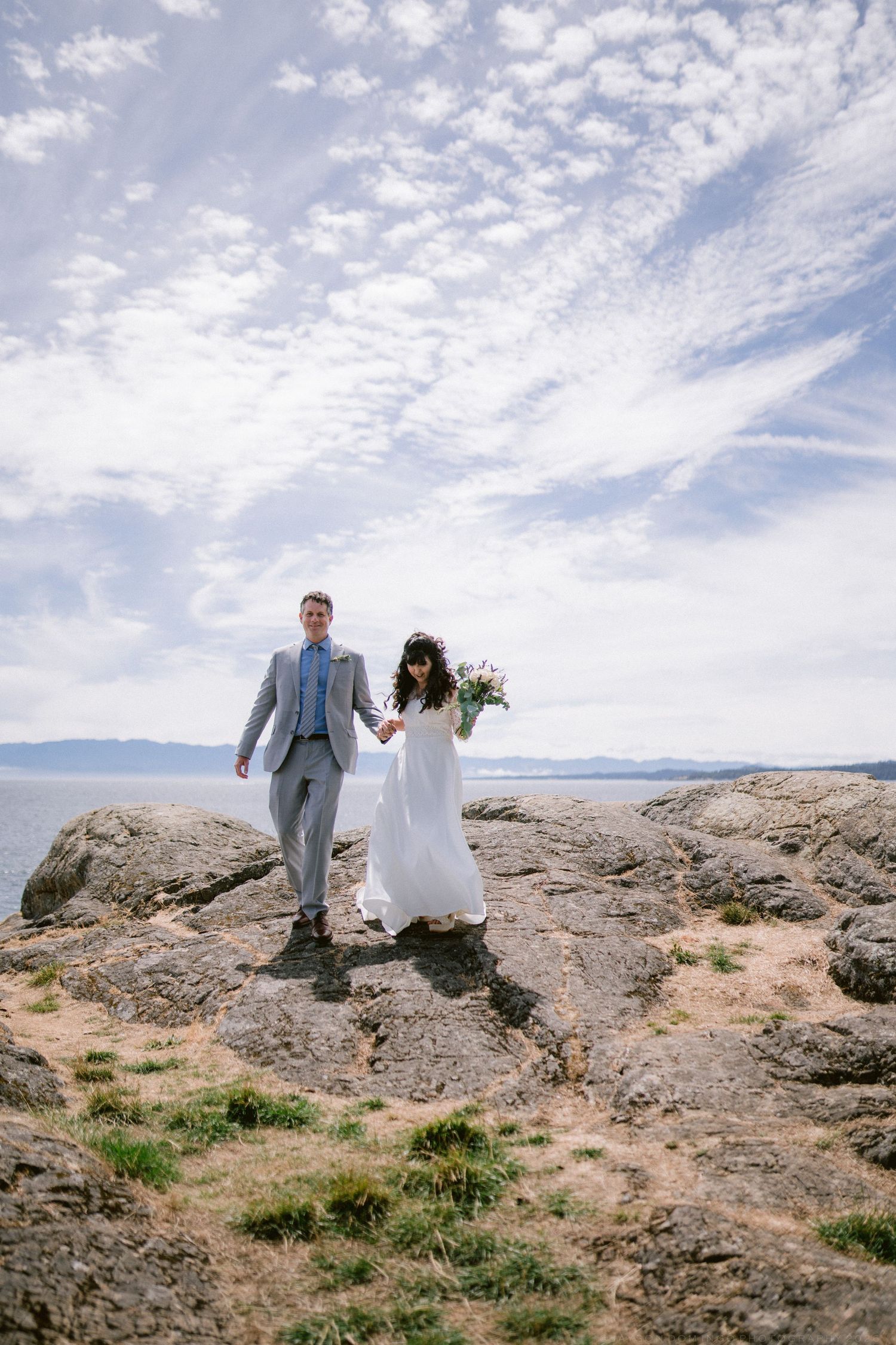 Bride and groom hold hands walking on rocky coastal cliffs under dramatic cloudy sky.