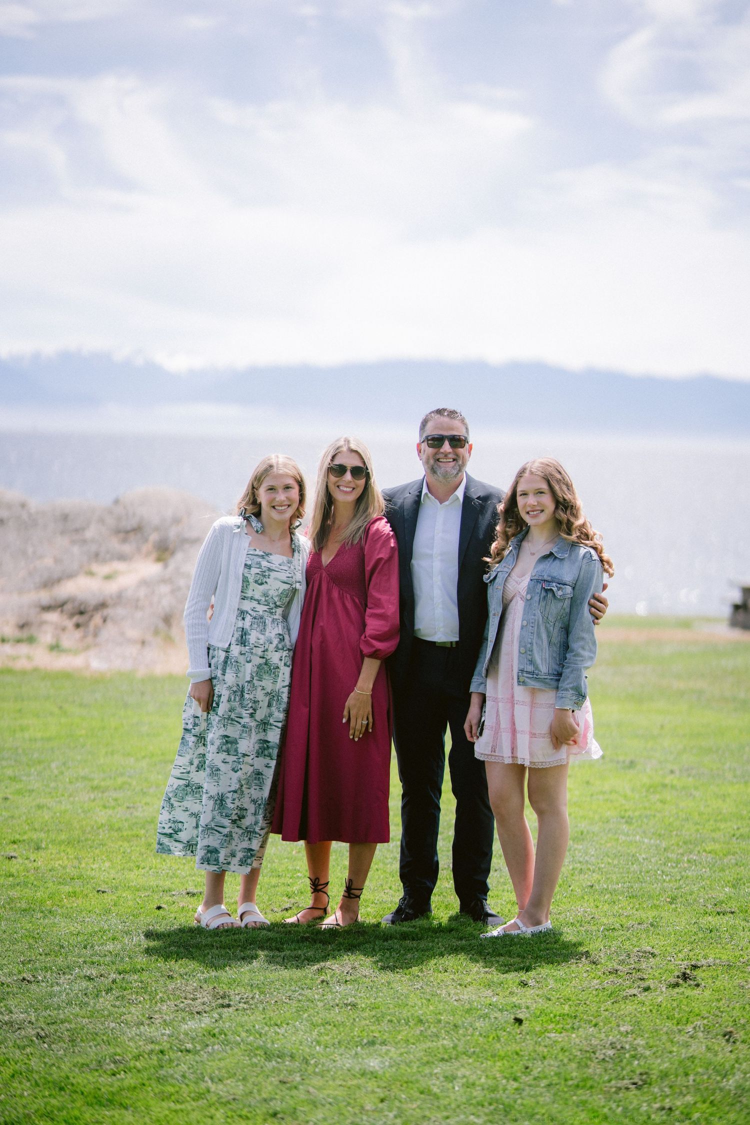 Family of four smiling together on green lawn with ocean and cloudy sky in the background.