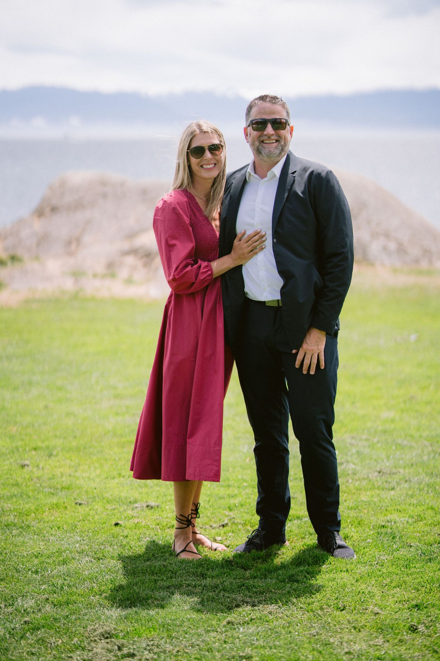 Smiling couple in sunglasses posing on green lawn with ocean view, woman in red dress.