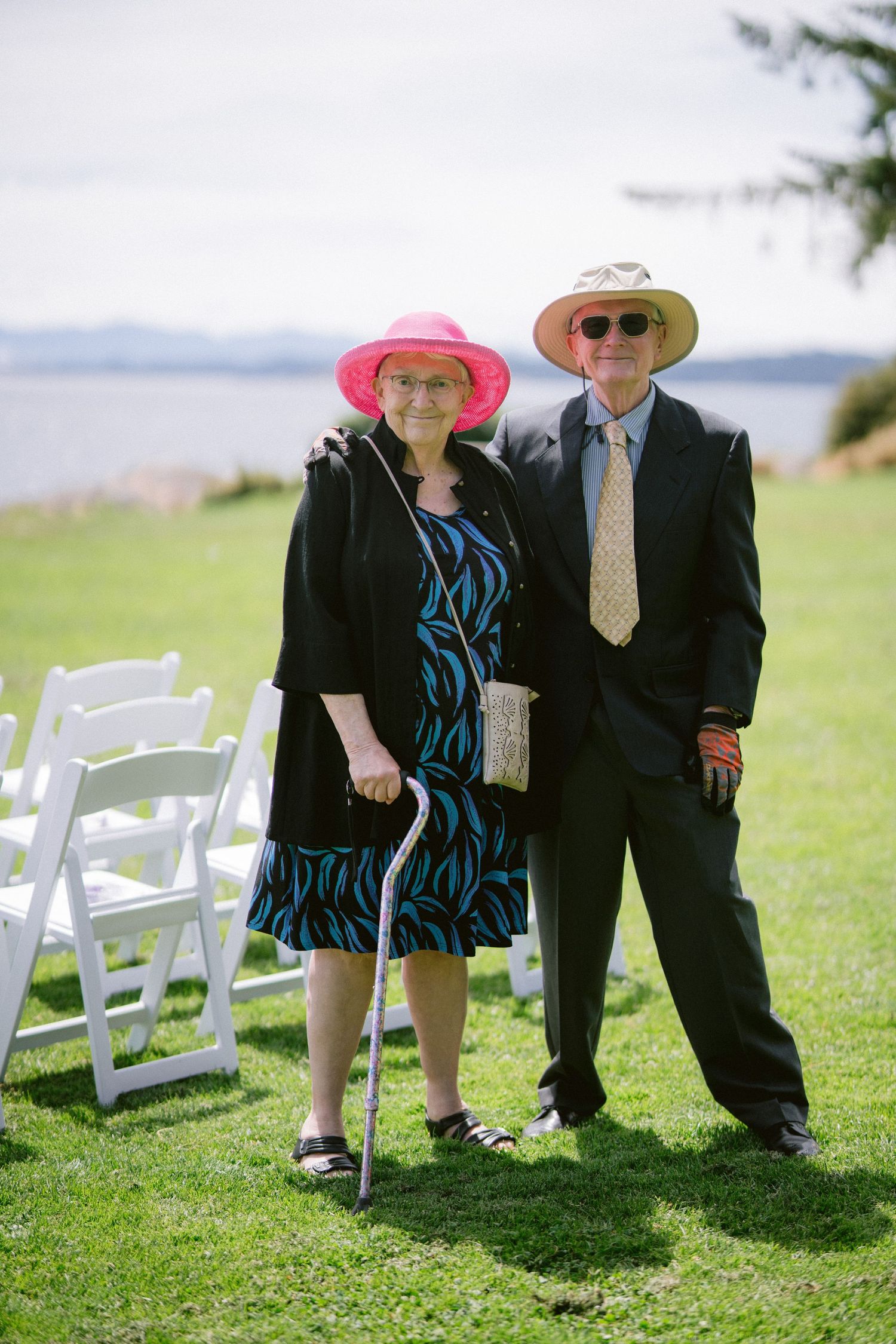 An elderly couple in formal attire and sun hats pose together on a lawn near white wedding chairs.