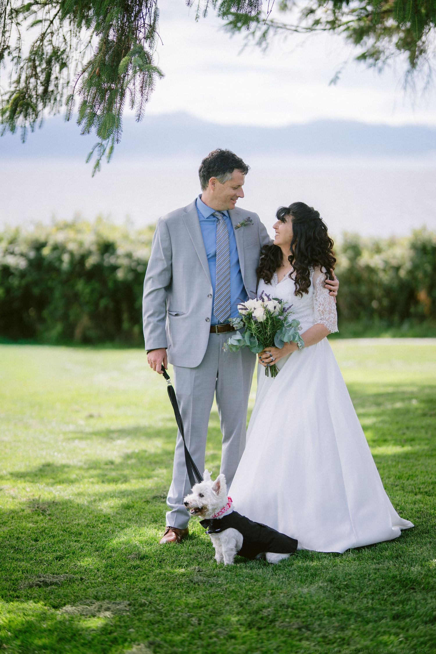 Bride and groom share a romantic moment on green lawn with small white dog on leash.
