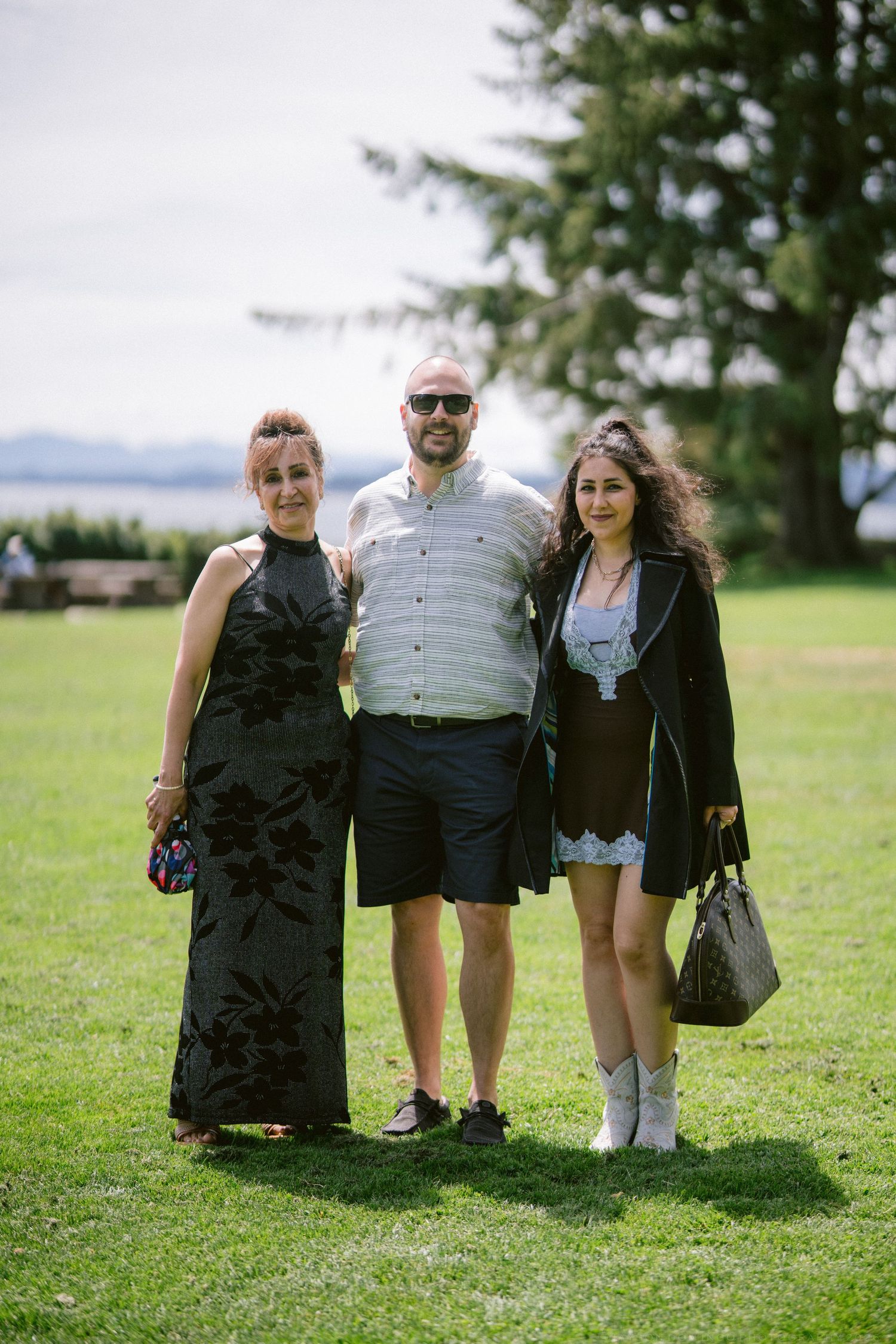 Three wedding guests posing on green lawn, man in shorts flanked by two women in formal attire.