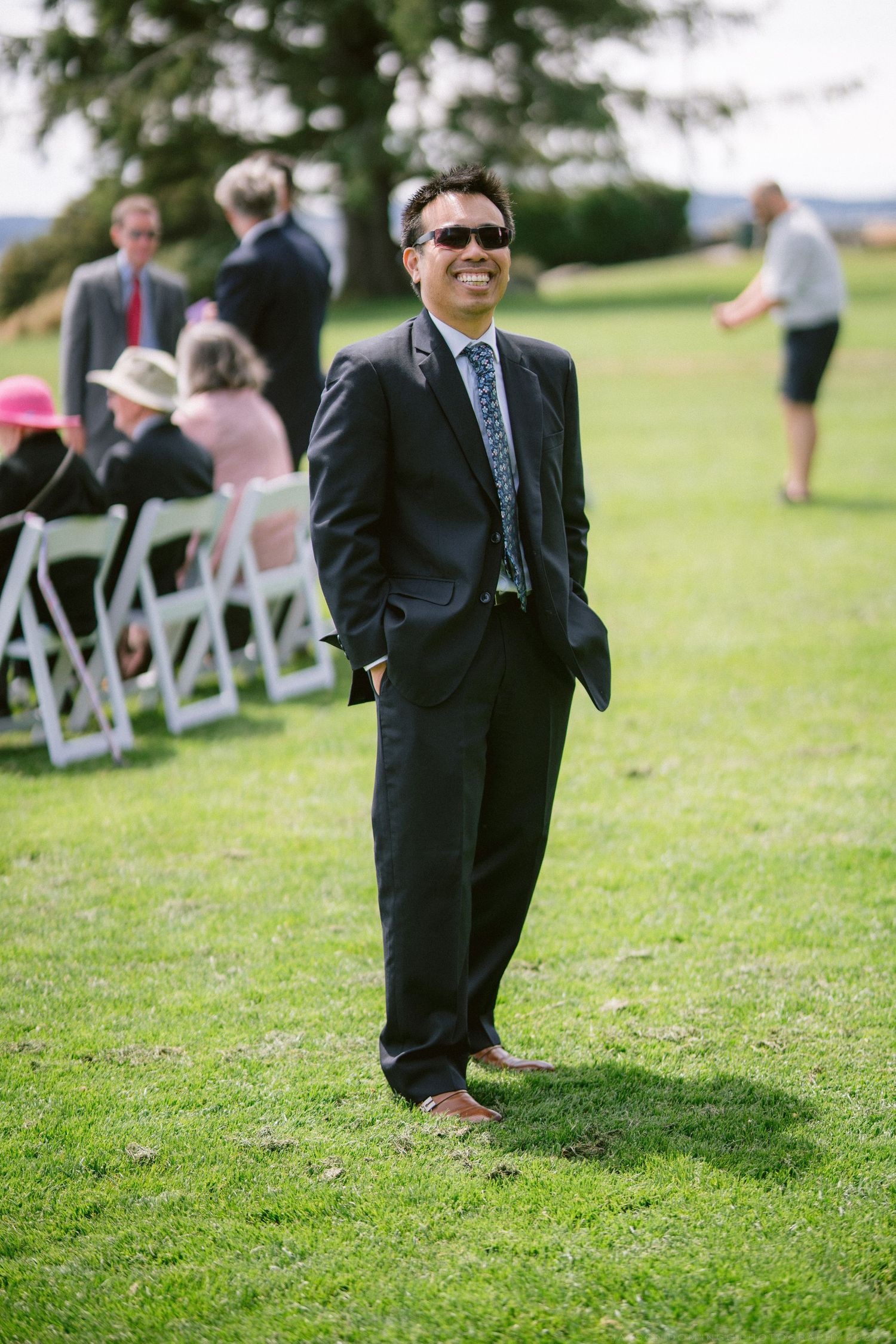Smiling man in black suit and sunglasses stands on green grass at outdoor wedding.