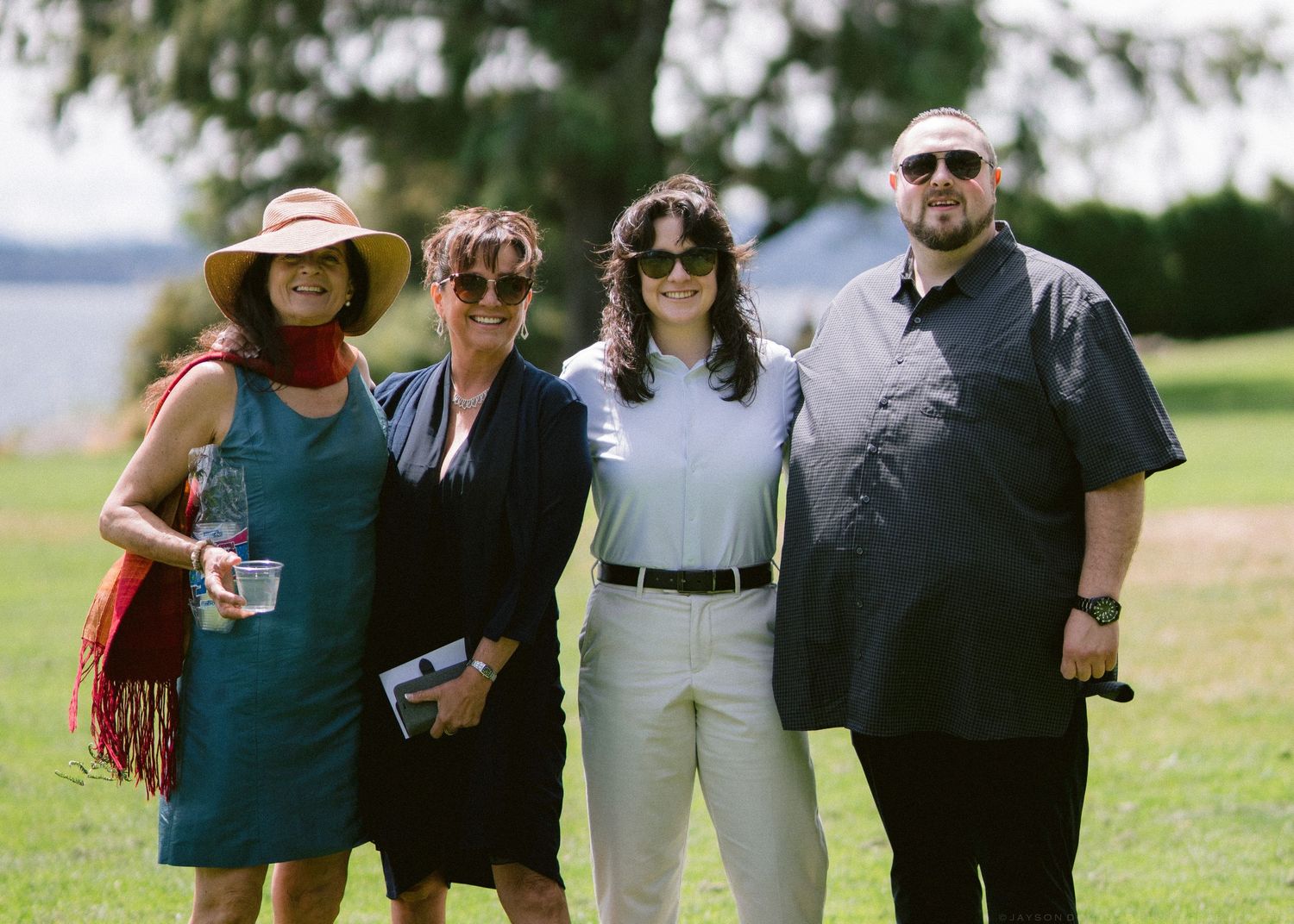 Four wedding guests smiling together on a sunny green lawn near a waterfront venue.