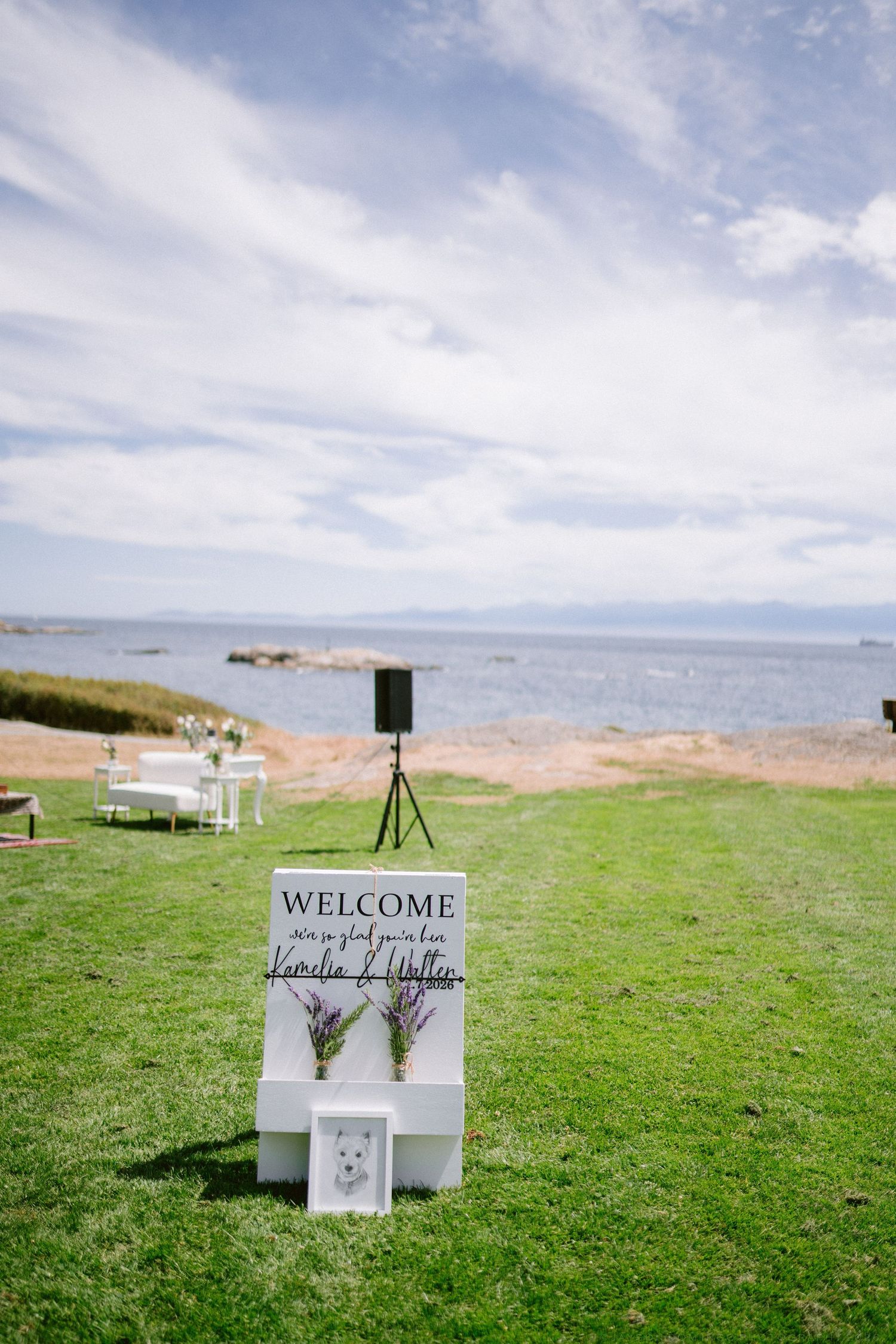 Outdoor wedding welcome sign on white stand with lavender, ocean backdrop, green lawn, and white reception tables.