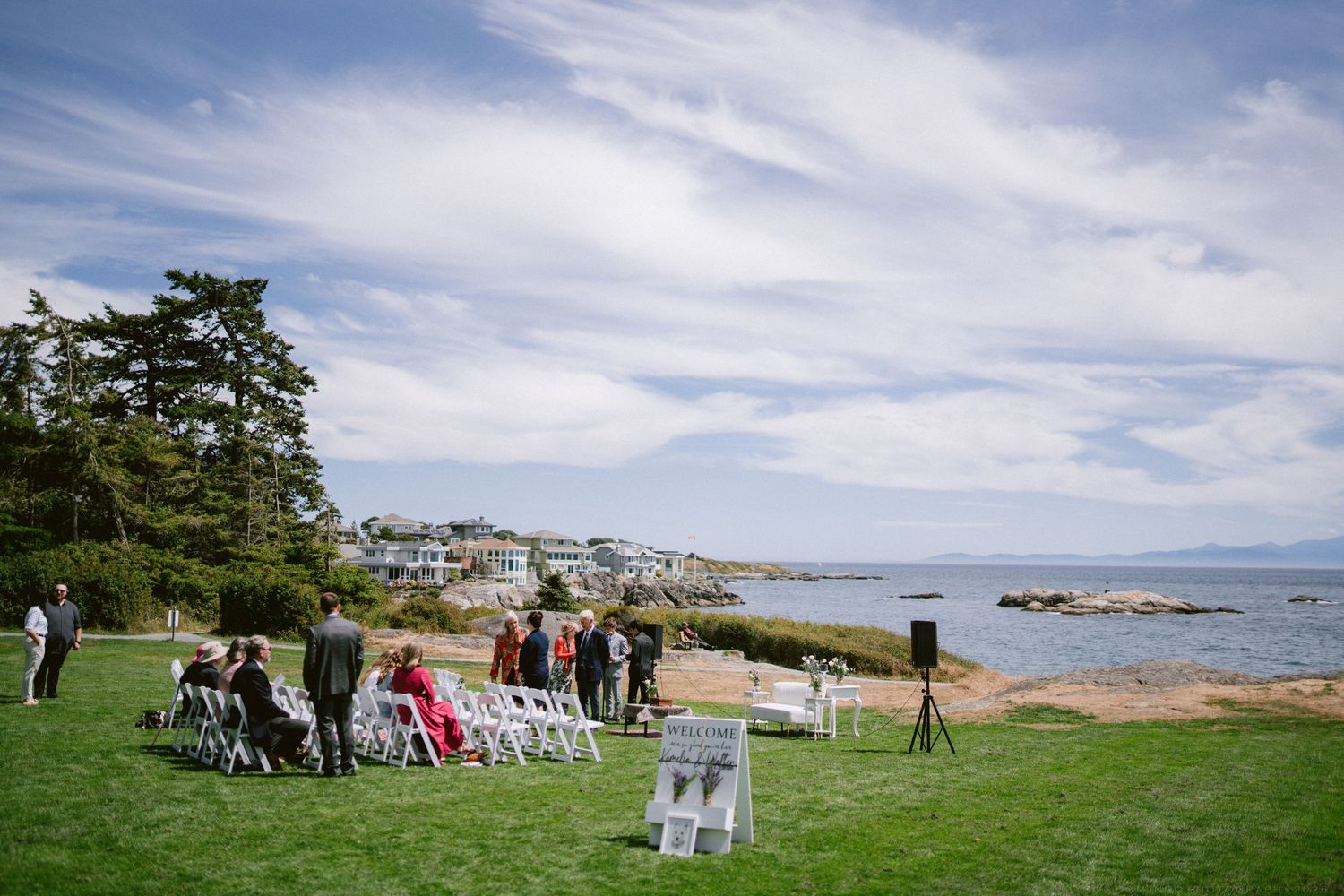 Outdoor seaside wedding ceremony with white chairs on green lawn overlooking rocky coastline.