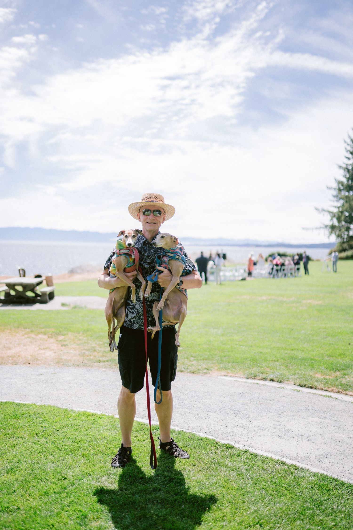 A smiling man in a straw hat holds two small dogs on leashes at an outdoor waterfront wedding venue.