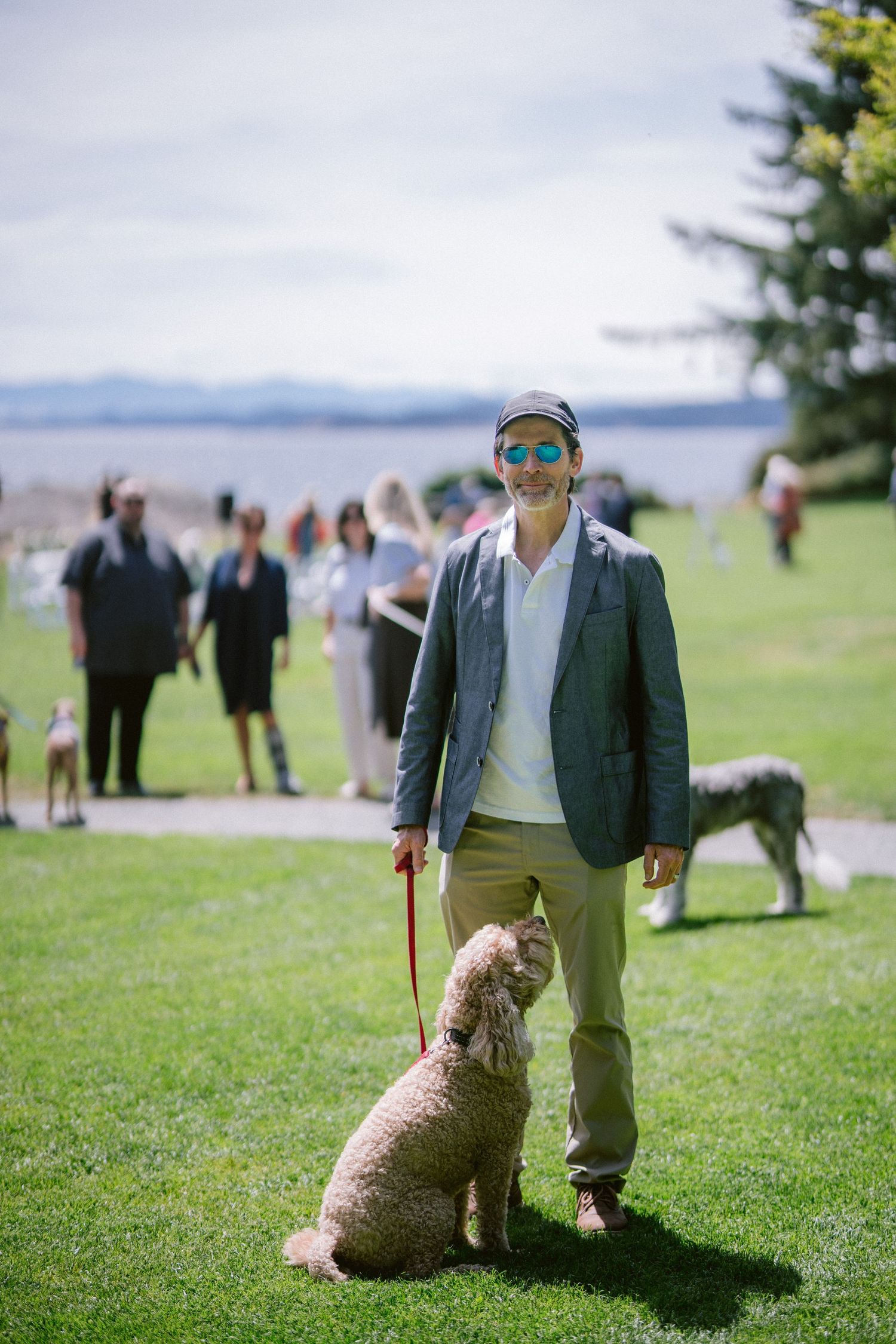 Man in blazer and sunglasses smiling with a goldendoodle on a leash in a sunny green park.