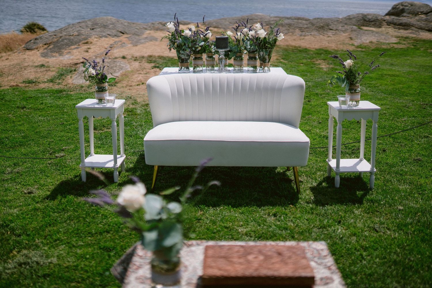 White tufted loveseat with floral arrangements on side tables set up for outdoor wedding reception.