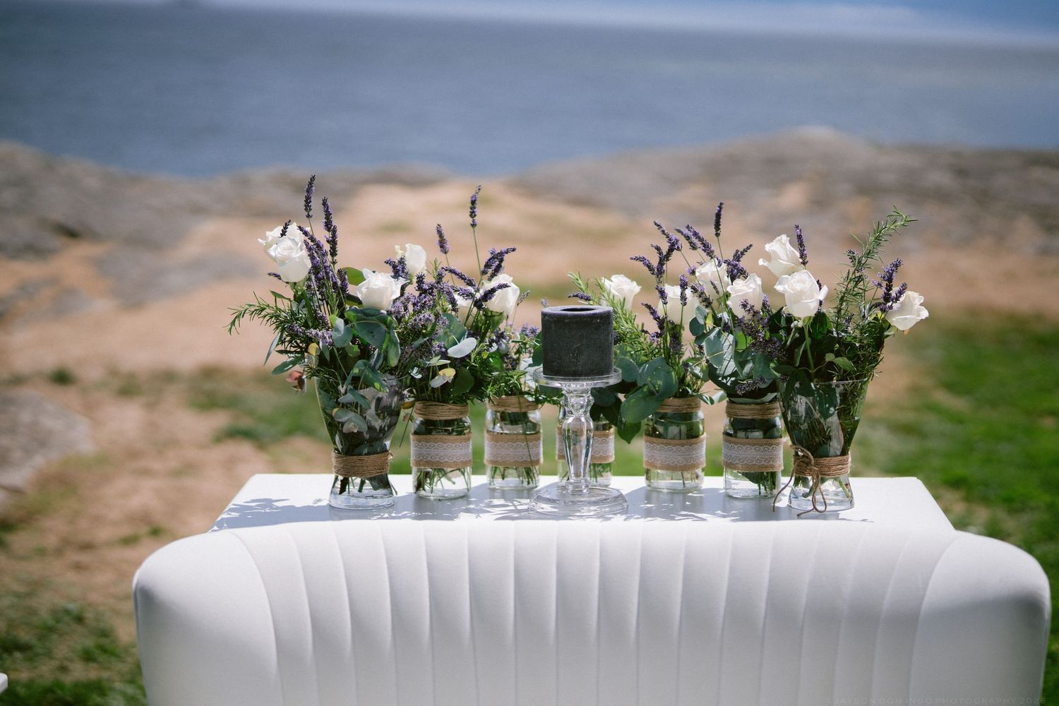 Elegant outdoor wedding table with white floral arrangements, lavender, and greenery set against a scenic coastal backdrop.