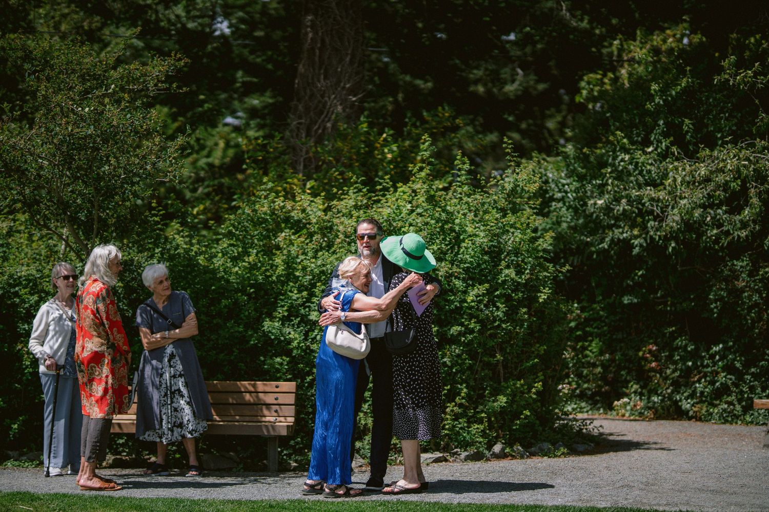 A group of wedding guests socialize and embrace on a shaded garden path surrounded by lush greenery.