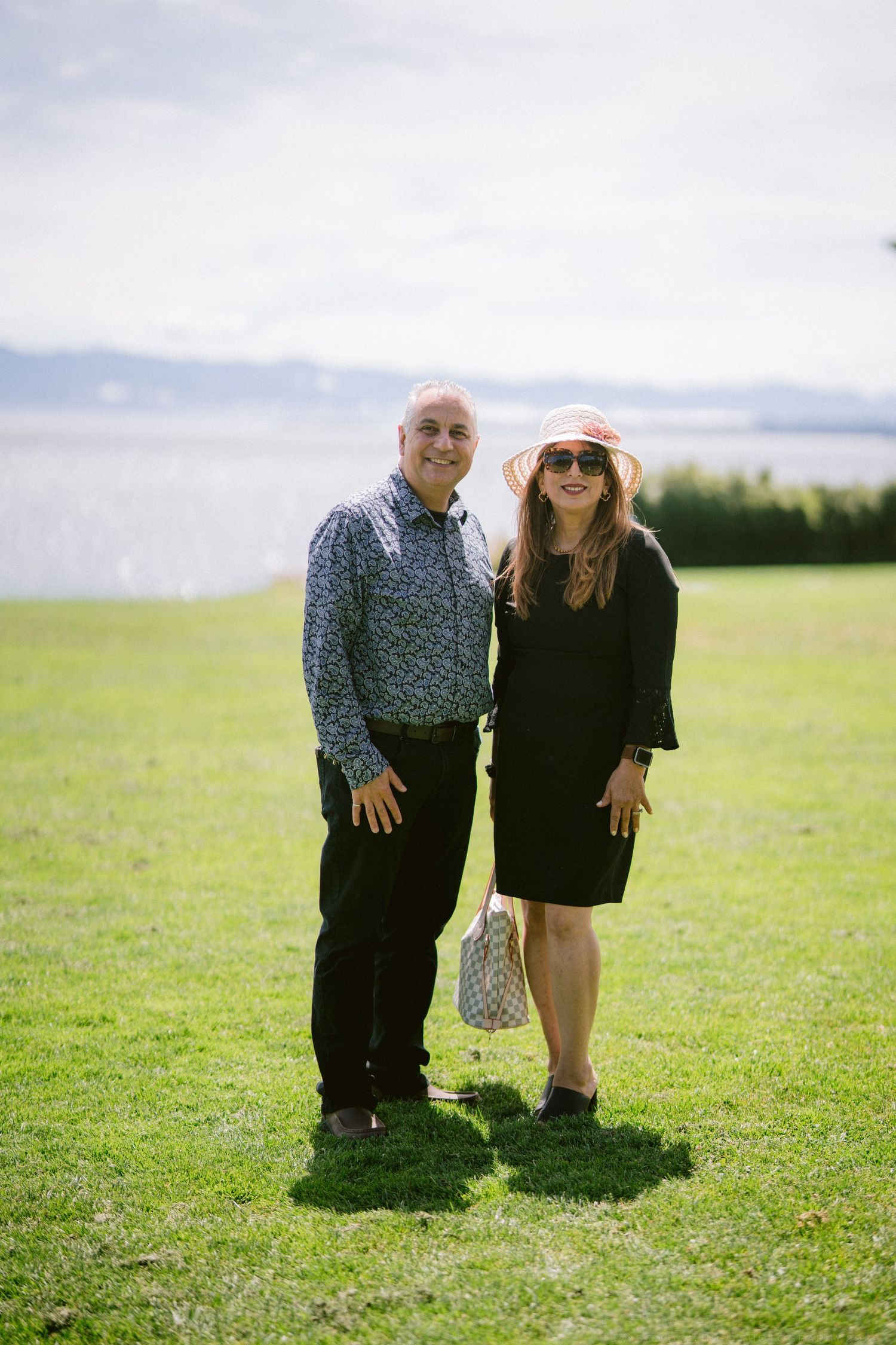 Smiling couple standing on green lawn at outdoor wedding with ocean and mountains in background.