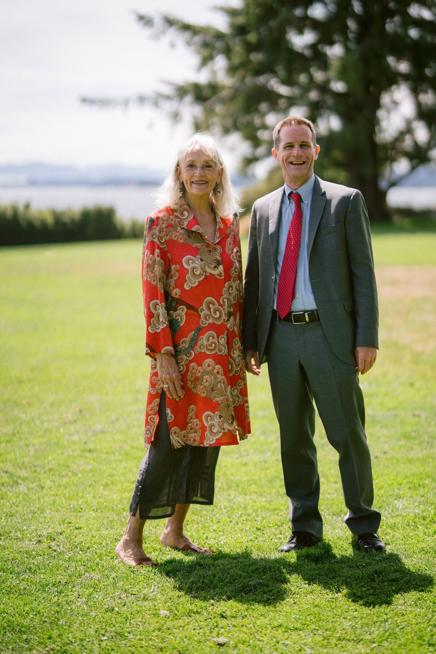 Smiling couple posing on green lawn, woman in red floral dress and man in gray suit with red tie.